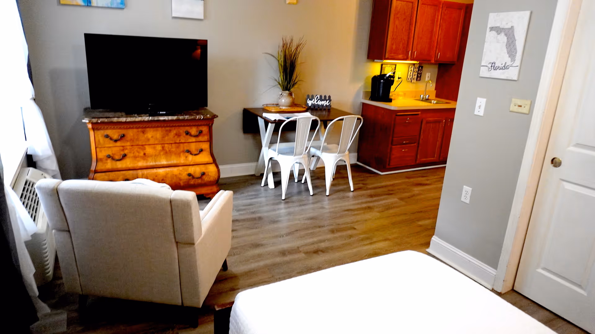 Studio-style room showing the corner of a bed, an armchair, a TV on a wooden dresser, a small dining table with chairs, and a kitchenette.