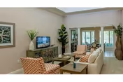 Bright living room with a sofa, two patterned armchairs, coffee table, TV on a cabinet, and plants by a sunlit doorway.