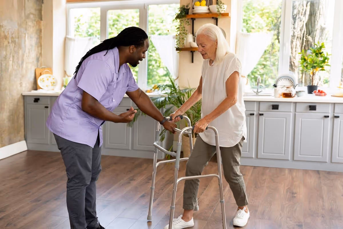 A caregiver in a purple shirt assists an elderly woman using a walker in a bright room with large windows and a kitchen area in the background.