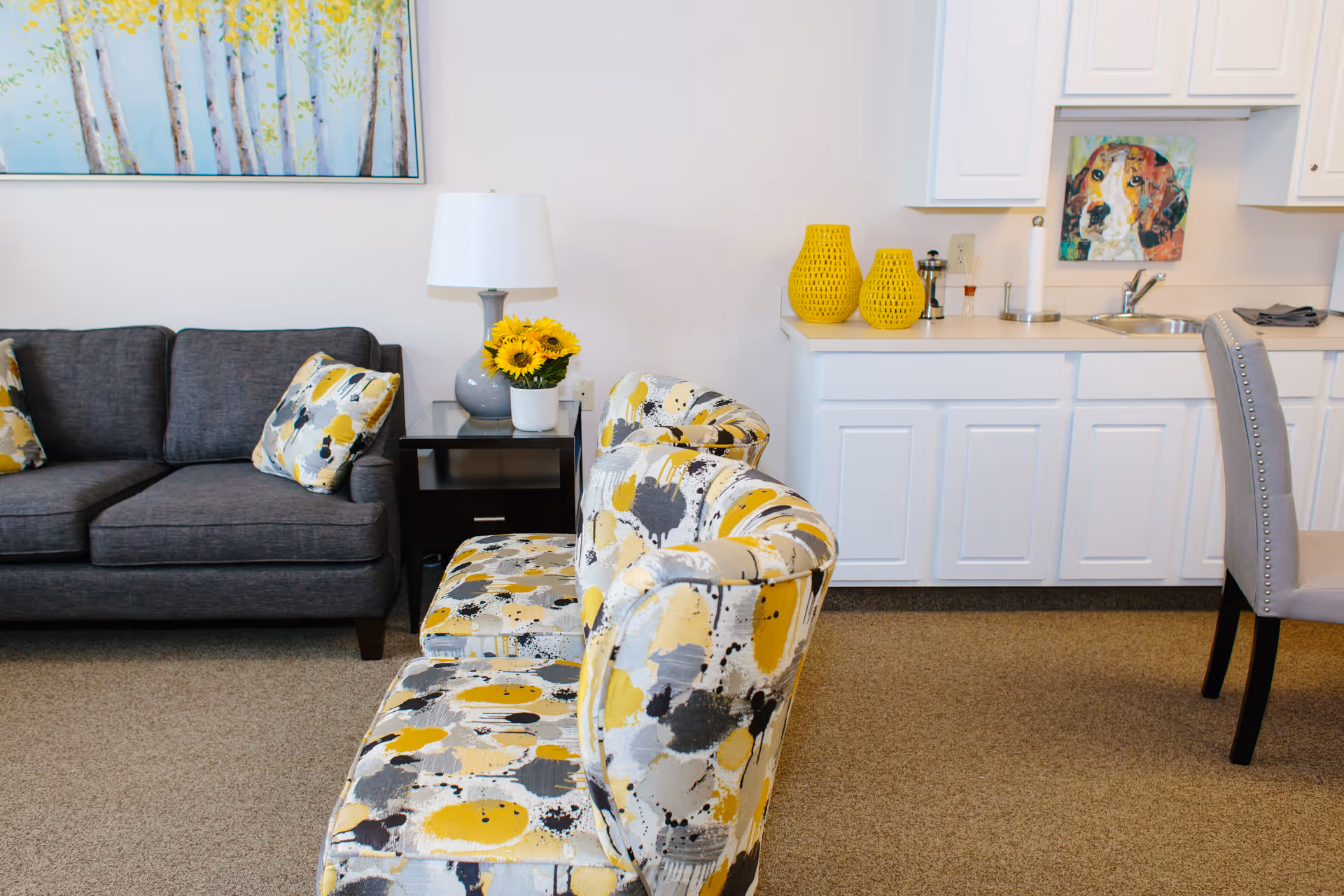 A cozy living area featuring a dark gray sofa with patterned yellow and gray pillows, two matching patterned chairs in yellow, gray, and white, a small black side table with a gray lamp and a vase of sunflowers, white cabinetry with a sink, and colorful artwork including a painting of a dog on the wall above the sink.