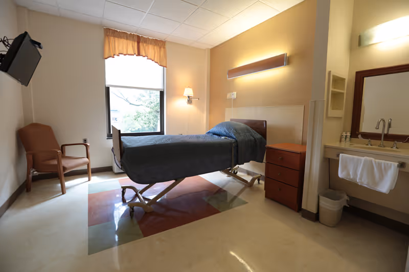 A simple, clean hospital-style bedroom with a single adjustable bed covered in a dark blue quilt. There is a wooden nightstand with drawers next to the bed, a brown armchair near a window with a beige curtain valance, and a sink with a mirror and towel on the right side. The room has beige walls and a tiled floor with a colored square pattern under the bed.