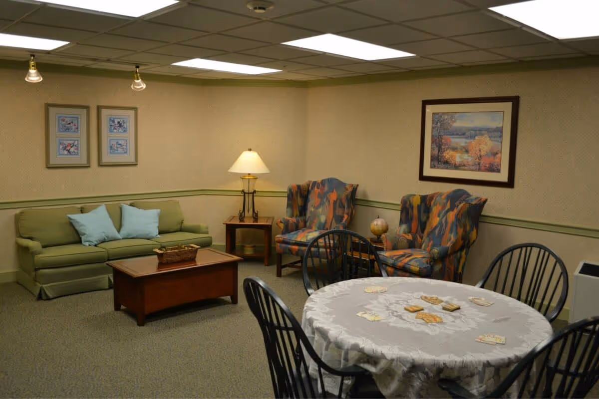 A cozy senior living common room with a green sofa adorned with two light blue pillows, a wooden coffee table with a basket, two colorful patterned armchairs, a side table with a lamp, and a round table covered with a white lace tablecloth set for a card game with cards and game pieces. The walls are decorated with framed artwork and the ceiling has fluorescent lighting.