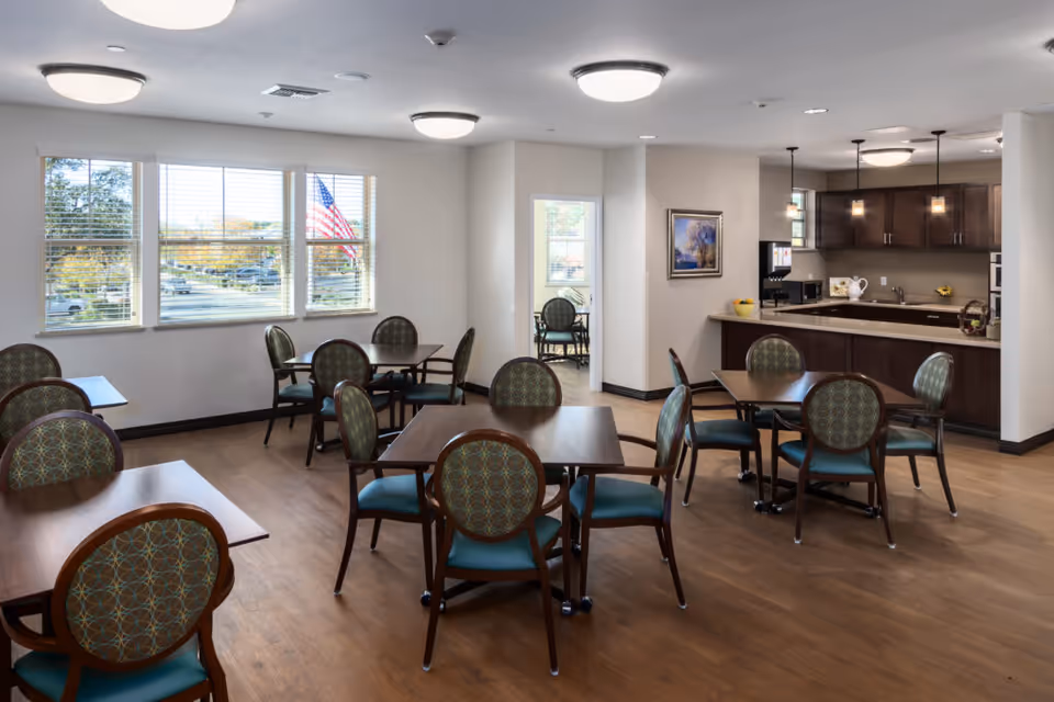 Well-lit dining area with multiple round tables and patterned chairs next to a kitchenette and large windows showing an American flag outside.