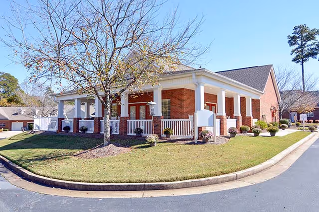 A single-story brick building with a white railing porch surrounded by a grassy lawn and a few small bushes and trees, under a clear blue sky.