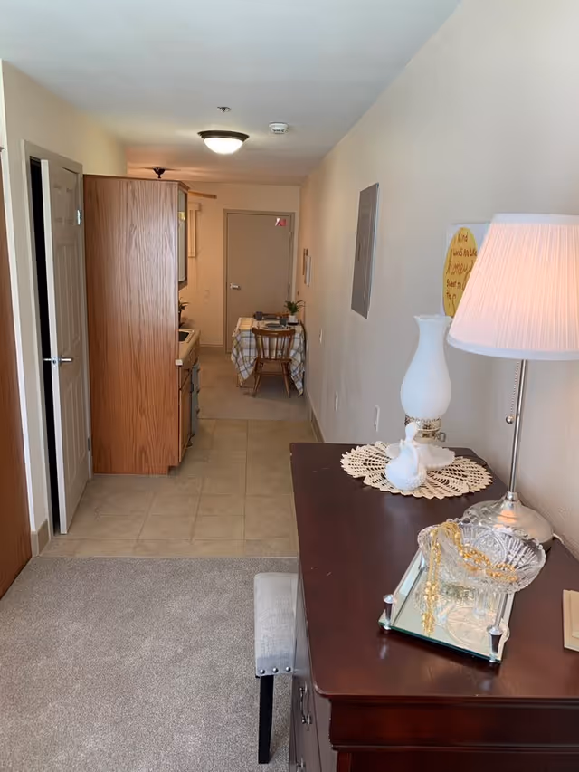 Interior view of a senior living facility apartment showing a narrow hallway with a small kitchen area on the left, a dining table with two chairs at the end, and a wooden dresser with a lamp, decorative items, and a cushioned stool on the right side in the foreground.