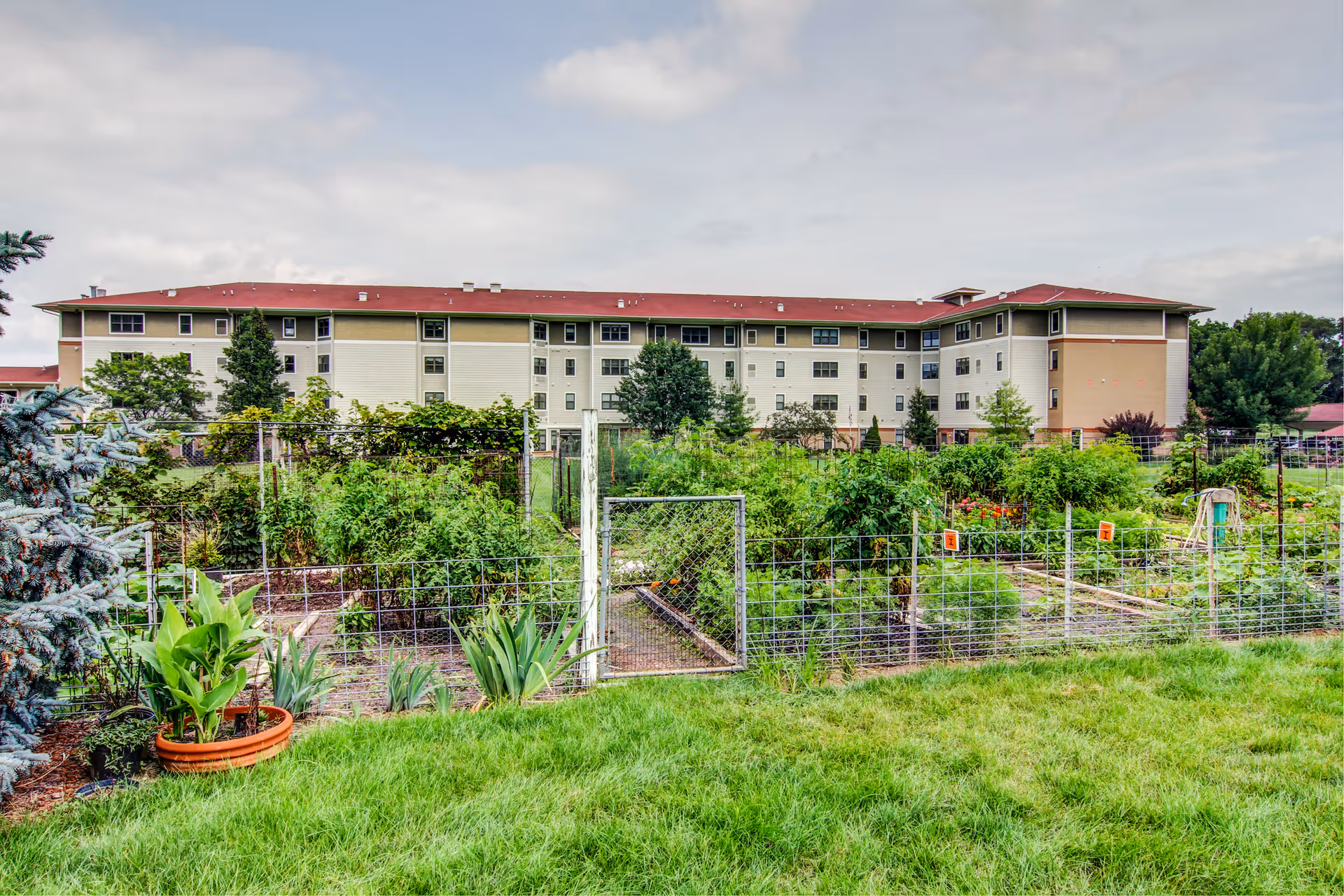 View of a multi-story residential building behind a fenced garden with various green plants and flowers under a cloudy sky.