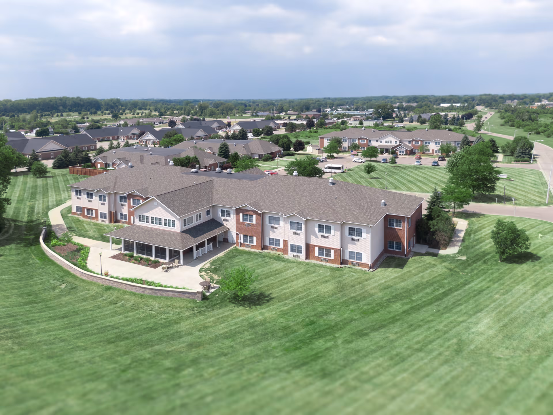 Aerial view of Charter Senior Living of Stepping Stone Falls showing multiple two-story buildings with brown roofs surrounded by well-maintained green lawns and trees under a partly cloudy sky.