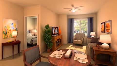 A cozy living and dining area in a senior living facility. The room features a round wooden dining table set for two with plates, glasses, and napkins. There is a comfortable armchair and sofa near a window with blue curtains, allowing natural light to brighten the space. A wooden TV stand holds a television and decorative items, and a green plant adds a touch of nature. The walls are adorned with colorful paintings, and a ceiling fan is mounted above. A doorway leads to a bedroom with a bed and bedside lamp visible.