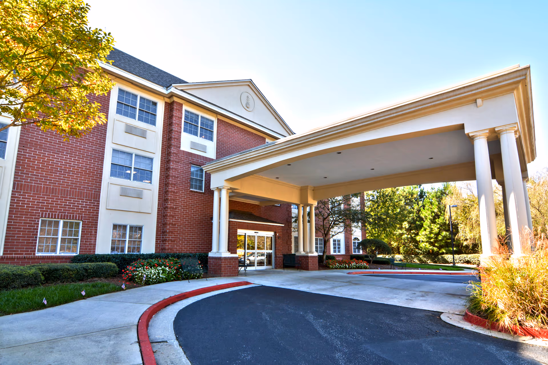 Exterior view of a senior living facility building with a covered entrance supported by white columns. The building has red brick walls with white trim around the windows. There are bushes and flowers planted along the walkway, and trees with green and yellow leaves are visible in the background under a clear blue sky.