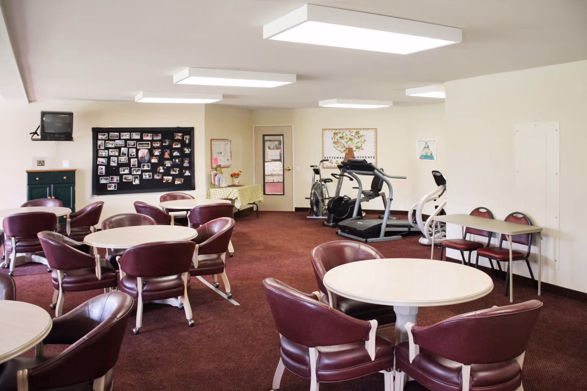 A common room with several round tables surrounded by maroon chairs on a carpeted floor. In the background, there is exercise equipment including a treadmill and stationary bike. The walls are light-colored with bulletin boards and a small TV mounted in the corner.