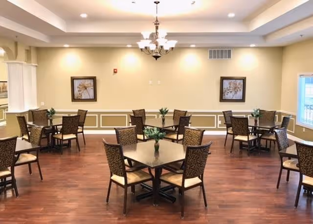 A well-lit dining room with several square tables, each surrounded by four patterned chairs. The room has wooden flooring, beige walls with white trim, two framed floral artworks, and a chandelier hanging from the ceiling. Small green plants are placed on each table.