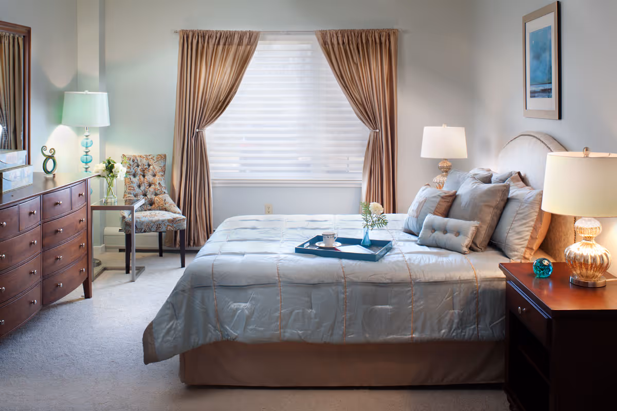 A well-lit bedroom with a large bed covered in a light blue comforter and multiple pillows. There is a wooden nightstand with a lamp and decorative items on the right side of the bed. On the left side of the room, there is a wooden dresser with a lamp and a vase of flowers, next to a patterned upholstered chair and a small side table. The window behind the bed has sheer blinds and brown curtains.