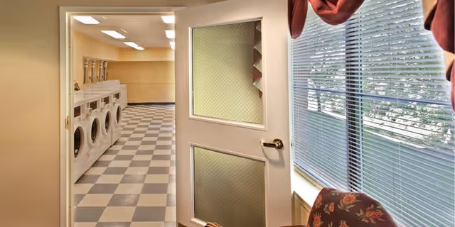 View through an open door into a laundry room with multiple front-loading washing machines lined up against the left wall. The floor has a checkered pattern of light and dark tiles. To the right of the door is a window with closed blinds and a floral-patterned chair partially visible.