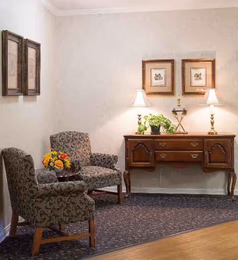 Cozy seating area with two patterned chairs, a wooden console table, lamps, plants, and framed art.