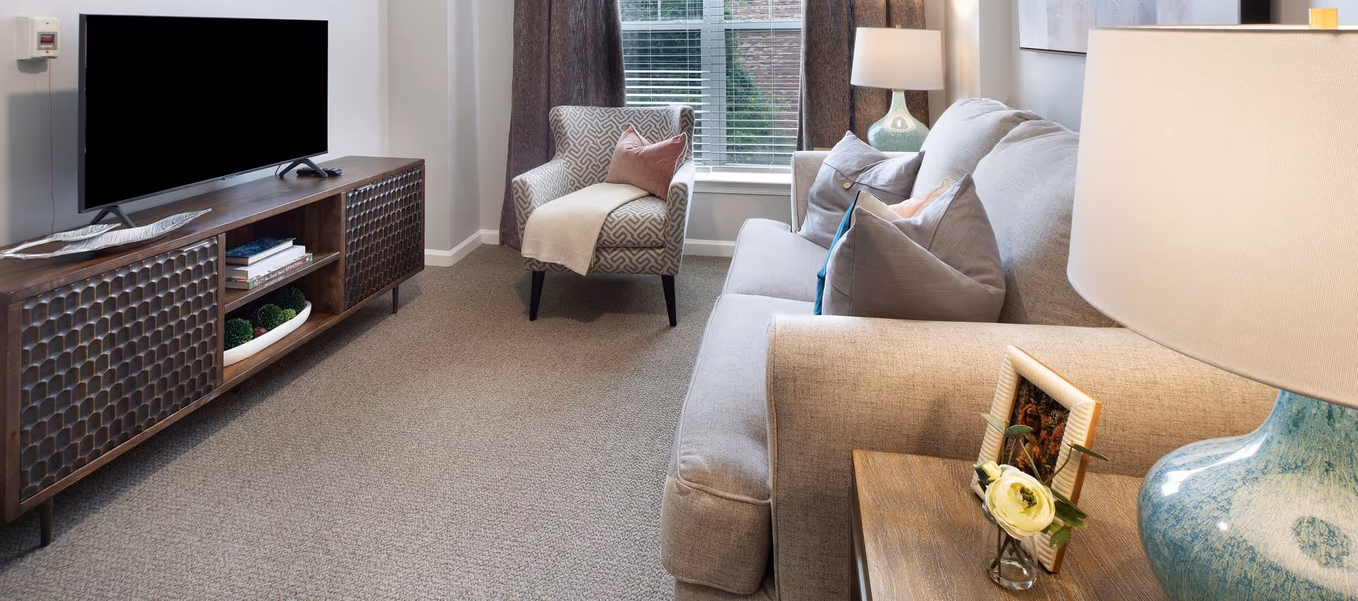 A cozy living room with a gray sofa adorned with multiple pillows, a patterned armchair with a throw blanket and pillow, a wooden TV stand with a flat-screen television, and a side table with a blue lamp, a small framed photo, and a flower in a glass vase. A window with blinds and curtains is in the background.