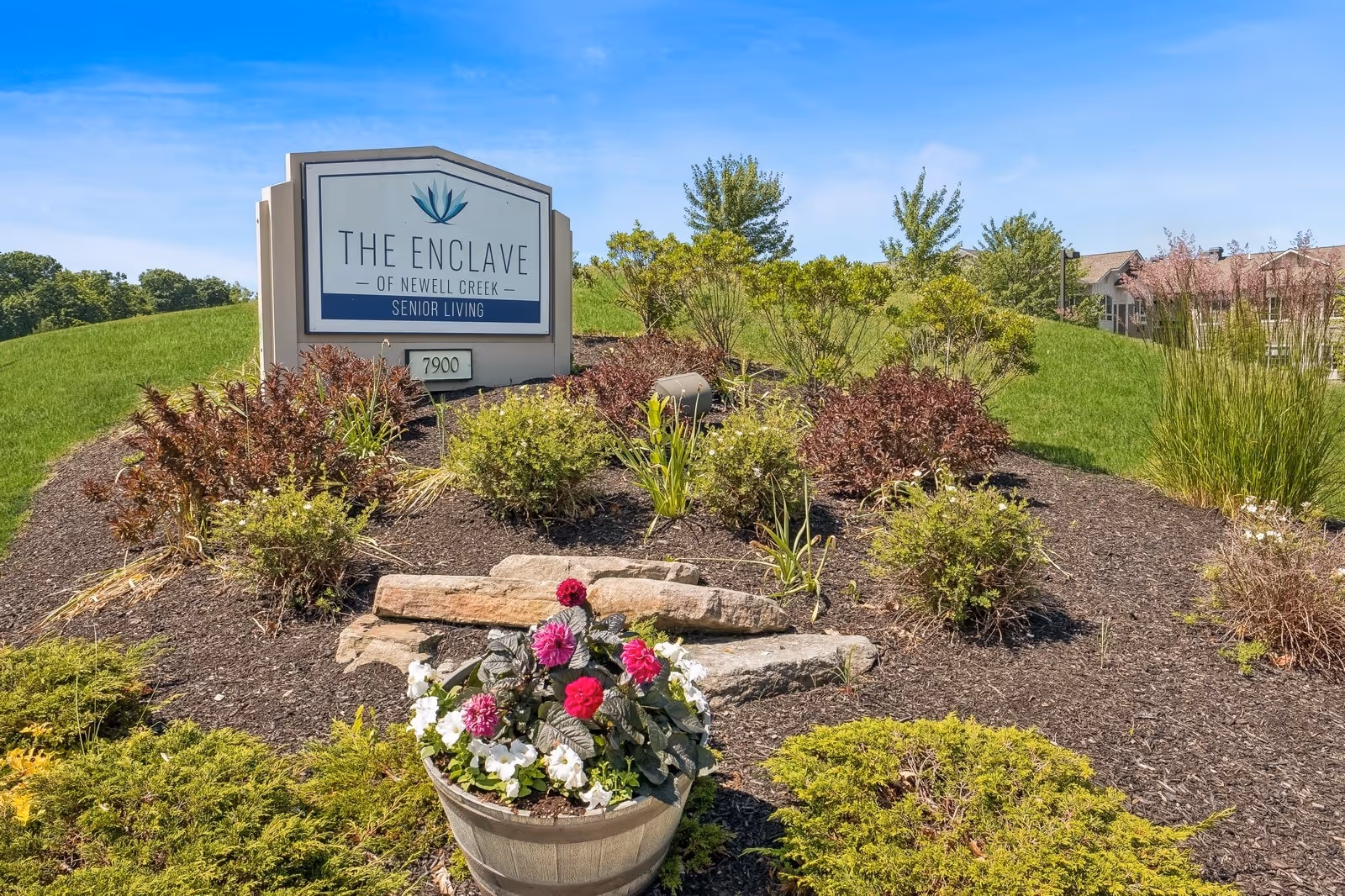 Outdoor landscaped area with a sign that reads 'The Enclave of Newell Creek Senior Living' surrounded by green grass, bushes, and a flower pot with pink and white flowers under a clear blue sky.