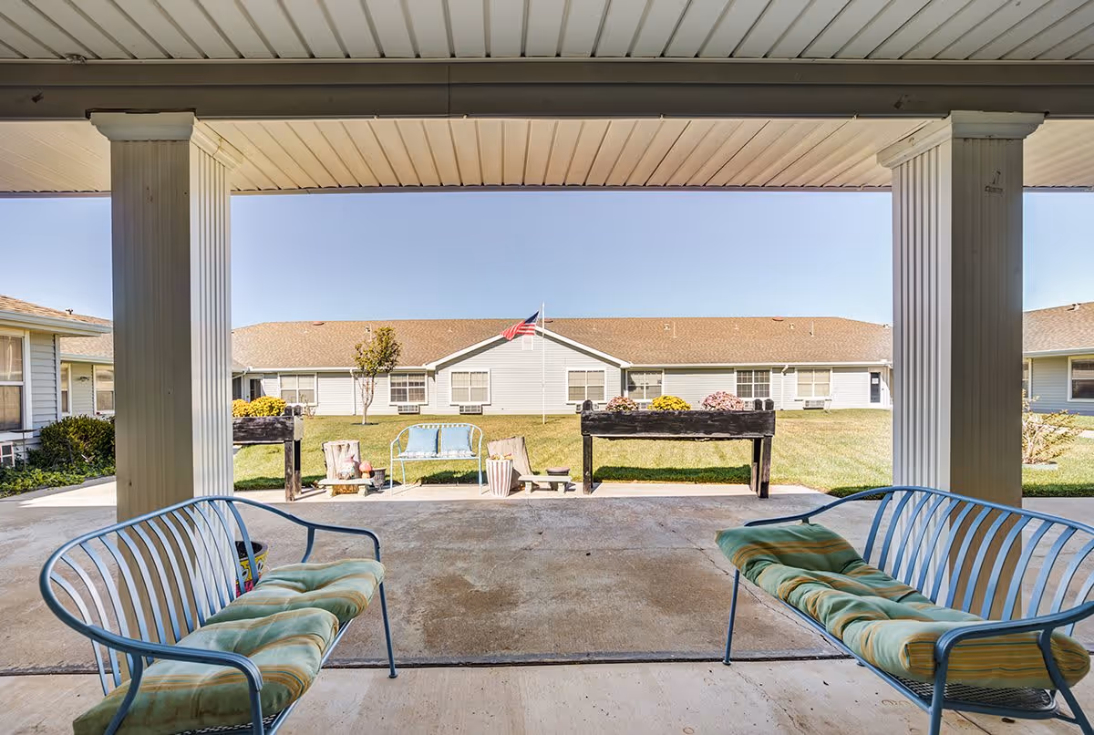 View from a covered patio area with two metal benches with cushions facing outward towards a grassy courtyard. In the courtyard, there are flower planters, a small blue bench, and an American flag on a flagpole in front of a single-story building with multiple windows.