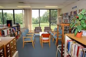 Small library/common room with bookshelves, chairs arranged around a table and large windows overlooking a green lawn.