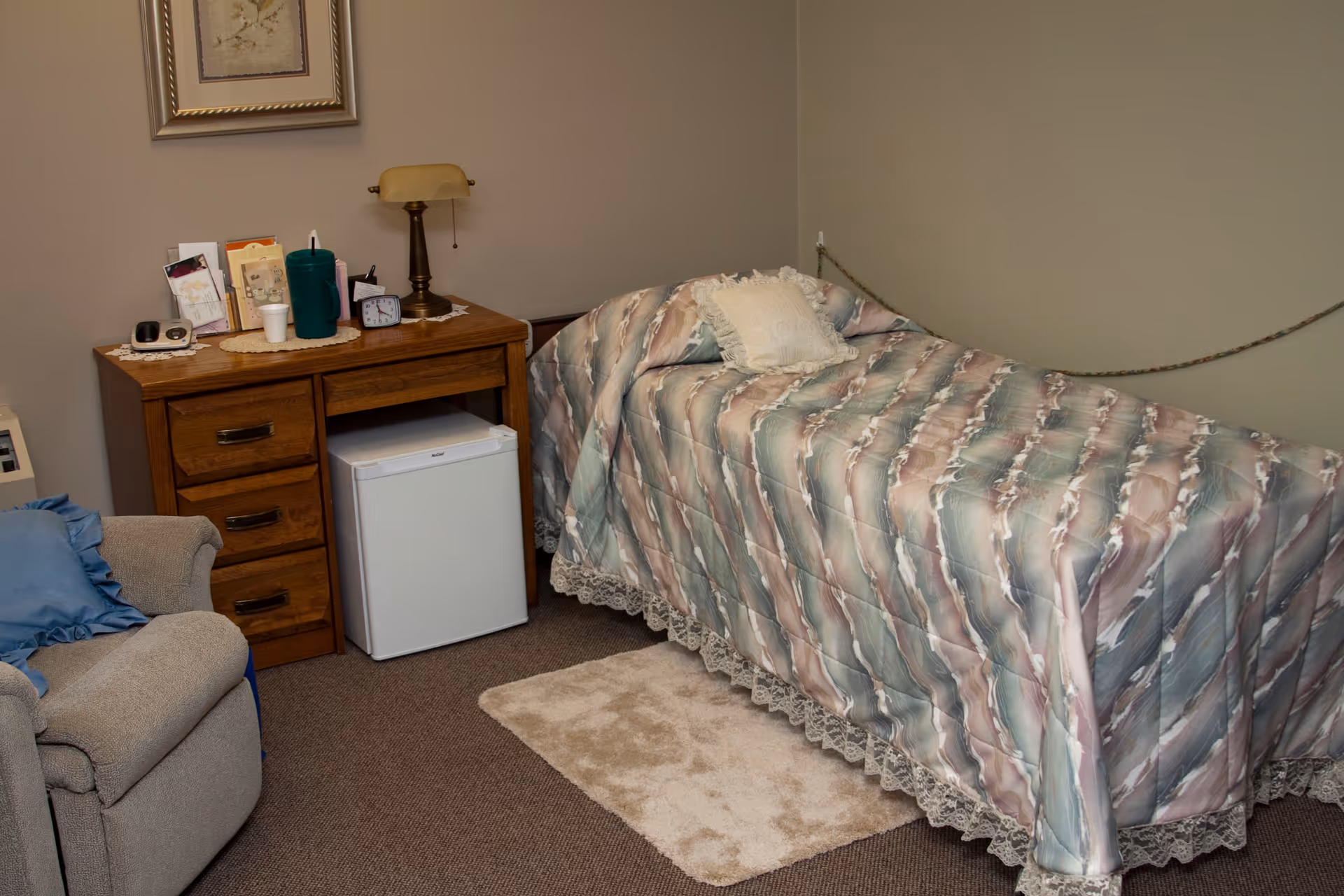 A small bedroom with a single bed covered in a patterned bedspread with a decorative pillow. Next to the bed is a wooden desk with drawers, a small white mini fridge underneath, a lamp, clock, and various small items on top. A beige armchair with a blue cushion is partially visible on the left side. The walls are plain and light-colored, with a framed picture hanging above the desk.