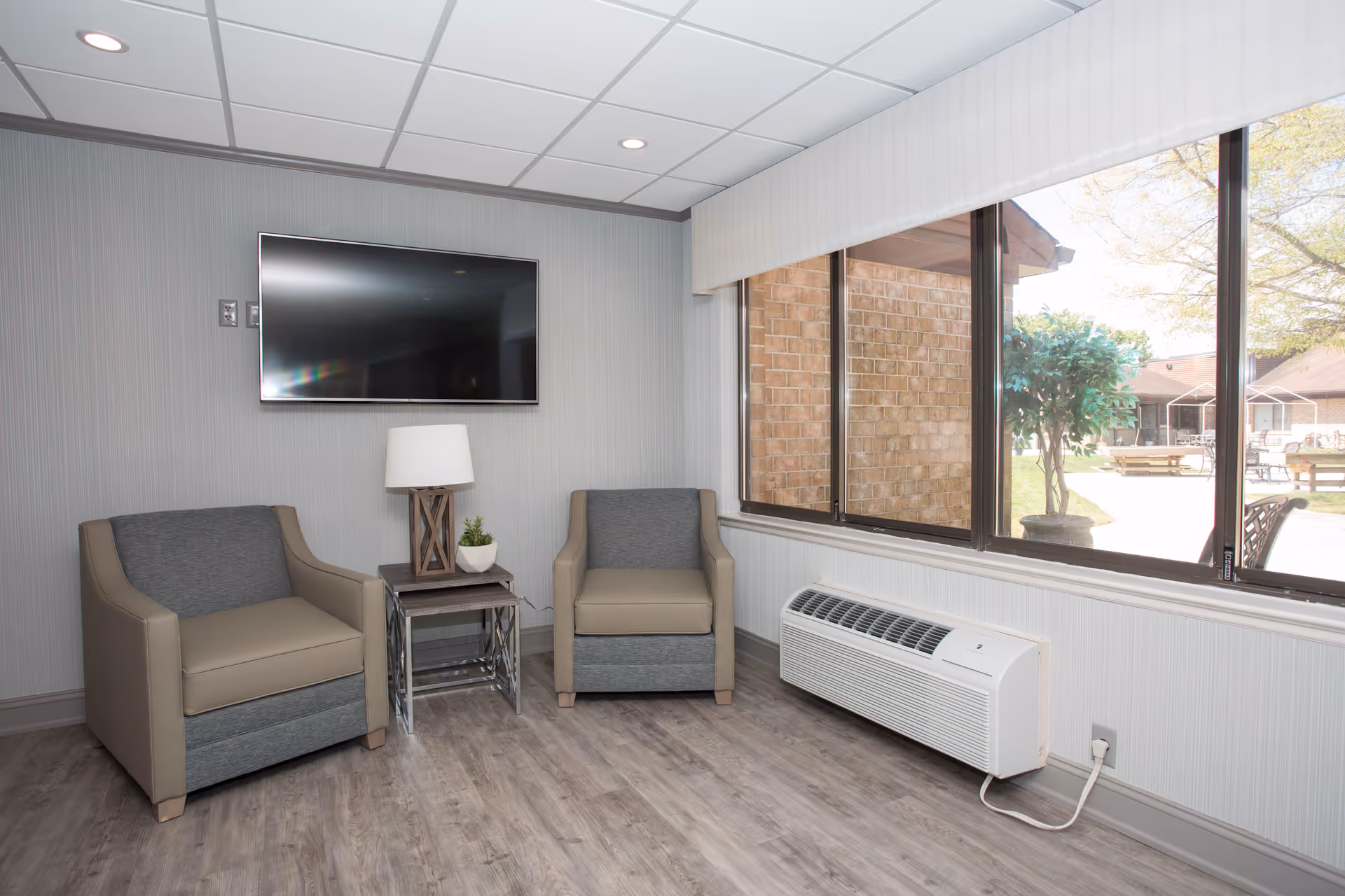 A small sitting area with two armchairs separated by a side table holding a lamp and a small plant. A flat-screen TV is mounted on the wall above the table. Large windows on the right side show an outdoor courtyard with trees and benches. The room has wood-look flooring and a wall-mounted air conditioning unit below the windows.