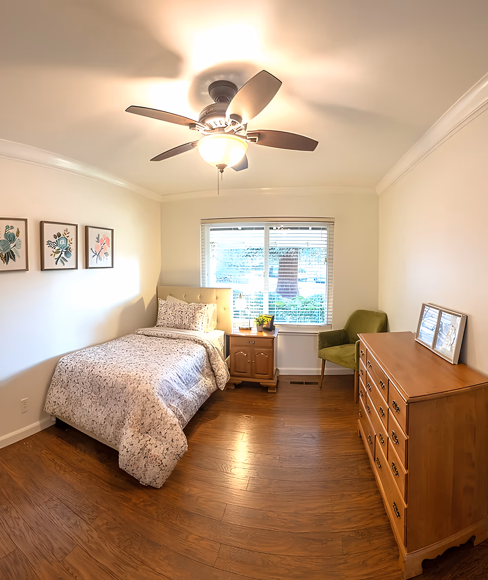 A cozy bedroom with a single bed covered in a patterned comforter, a wooden nightstand with a small plant and a book, a green upholstered chair, a wooden dresser with framed pictures on top, three framed floral artworks on the wall, a ceiling fan with a light, and a window with blinds letting in natural light.