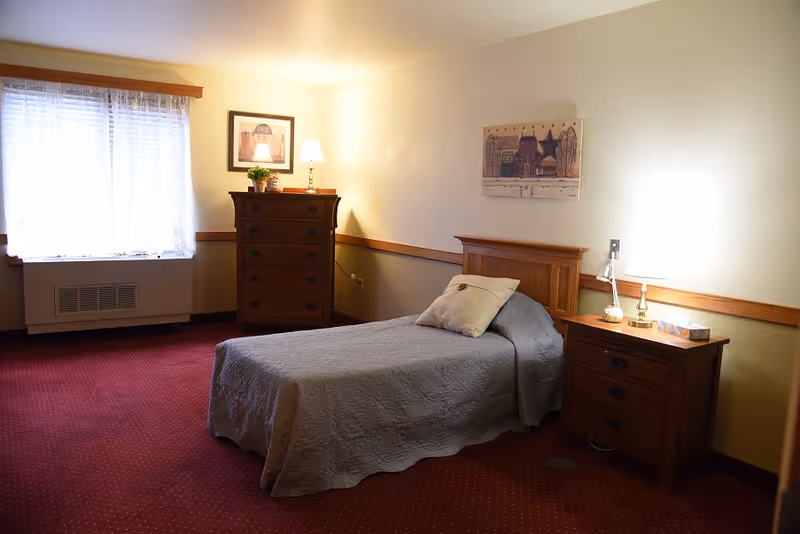 A cozy senior living bedroom with a single bed covered in a gray quilt, a pillow, a wooden nightstand with a lamp and tissue box, a wooden dresser with a lamp and plant, a window with sheer curtains, and framed artwork on the walls.