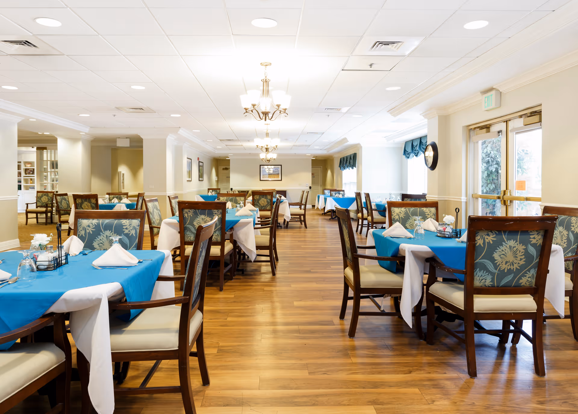 A bright and spacious dining room with wooden floors, multiple tables covered with white and blue tablecloths, and chairs with floral patterned cushions. The room is well-lit with ceiling lights and chandeliers, and there are large windows with blue valances letting in natural light. The walls are painted a light color and decorated with framed artwork.