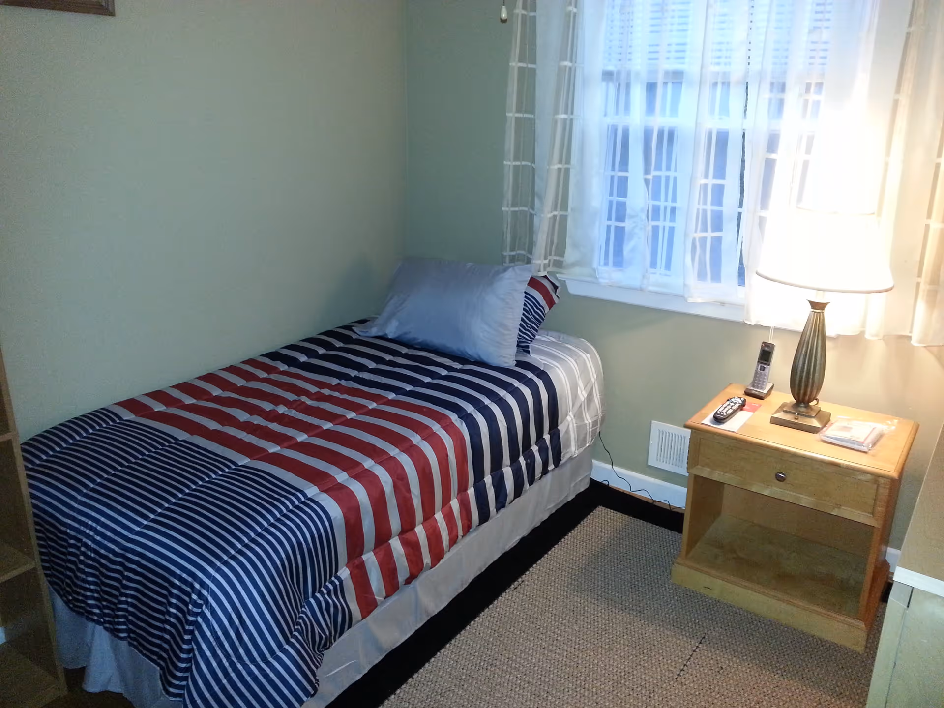 Small bedroom with a single bed covered in red, white, and blue striped bedding, a pillow, a window with sheer curtains, and a wooden nightstand holding a lamp and phone.