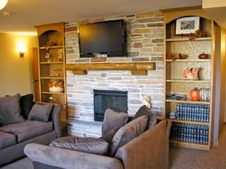 Cozy living room with a stone fireplace in the center, a flat-screen TV mounted above the mantel, built-in wooden shelves on either side displaying books and decorative items, and two brown upholstered sofas facing the fireplace.