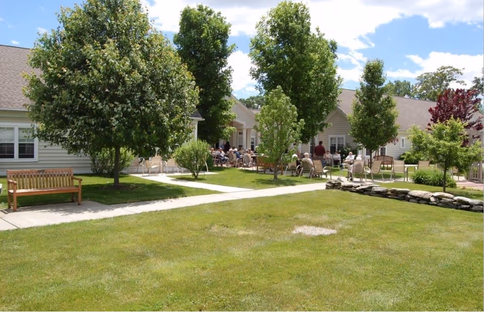 Outdoor courtyard area of Yorktown Assisted Living Residence with green grass, several trees, wooden benches, and multiple people sitting on chairs near the building under a partly cloudy sky.