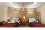 A simple, clean room with two single beds separated by a wooden nightstand. Each bed has a burgundy blanket and a pillow with a floral design. There are wall-mounted reading lights above each bed and a small lamp on the nightstand. The room has beige walls and a curtain on the right side.