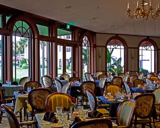 Elegant dining room with multiple tables set with glassware, plates, and napkins. The room features large arched windows with wooden frames allowing natural light to enter, and upholstered chairs with floral and striped patterns. A chandelier hangs from the ceiling, and greenery is visible outside the windows.