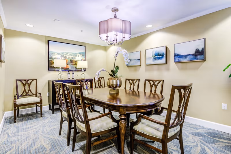 A well-lit dining room with a wooden oval dining table surrounded by eight matching chairs with cushioned seats. A decorative plant in a gold vase is centered on the table. The walls are painted light yellow and adorned with four framed landscape paintings. Two table lamps with glass bases and beige shades sit on a sideboard against the far wall. A modern chandelier with a fabric shade hangs above the table. The floor is carpeted with a blue and beige patterned rug.