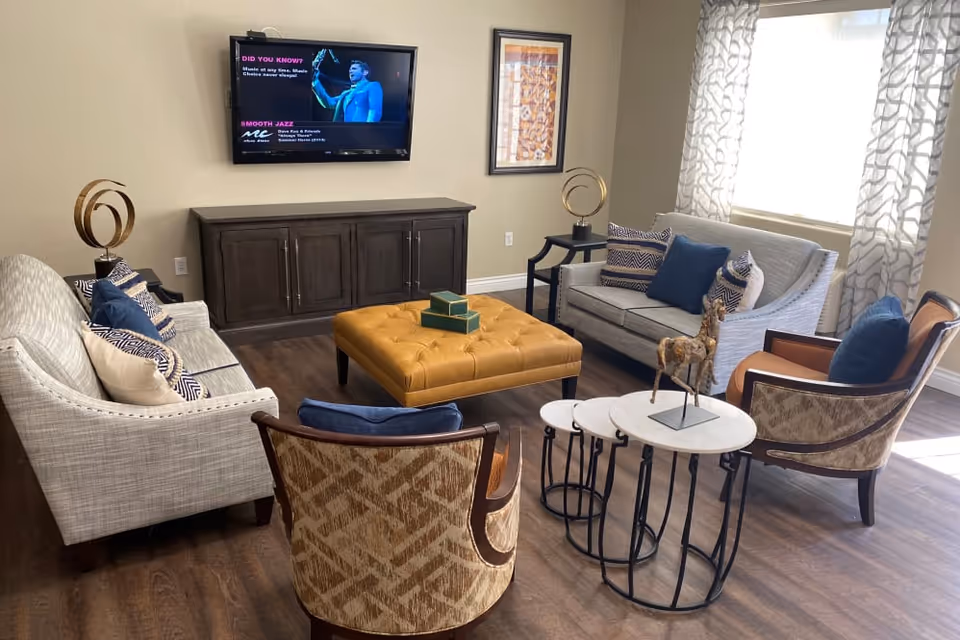 A cozy living room with two light gray sofas adorned with blue and patterned throw pillows, two patterned armchairs with blue cushions, a large mustard yellow tufted ottoman in the center, and two round nesting tables with a decorative horse sculpture. A flat-screen TV is mounted on the wall above a dark wooden cabinet, and a framed artwork hangs on the wall next to the TV. Light filters in through a window with patterned curtains.