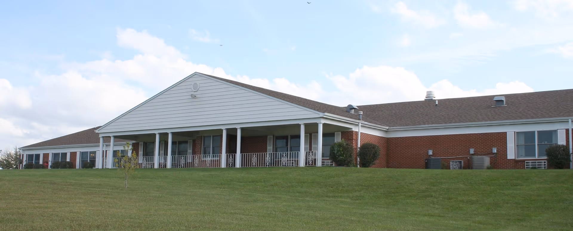 Low, single-story brick senior living building with a covered porch and columns on a grassy lawn under a partly cloudy sky.