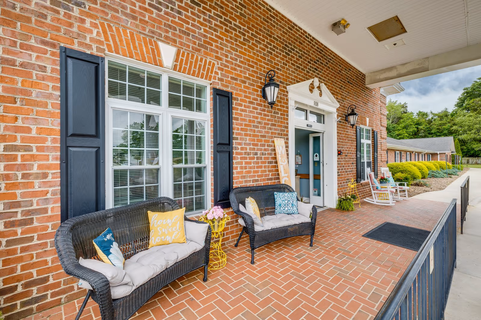 Covered brick front entrance with wicker benches, decorative pillows, and potted plants on a tiled porch.