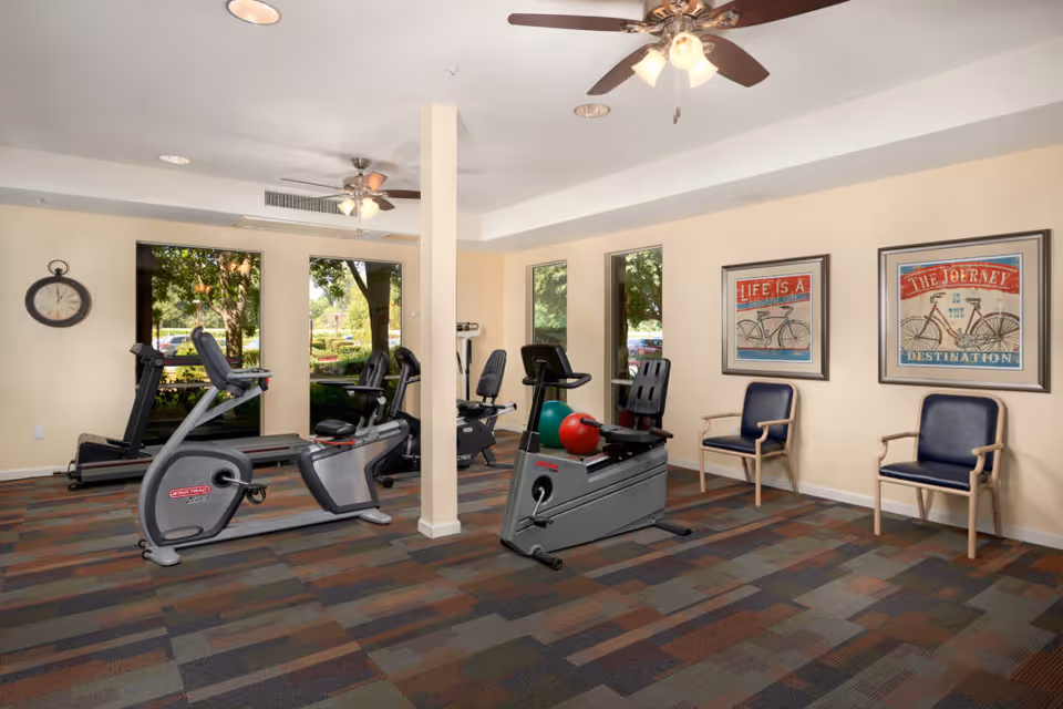 Bright interior exercise room with recumbent bikes, a treadmill, seating, exercise balls, and framed bicycle prints by large windows.
