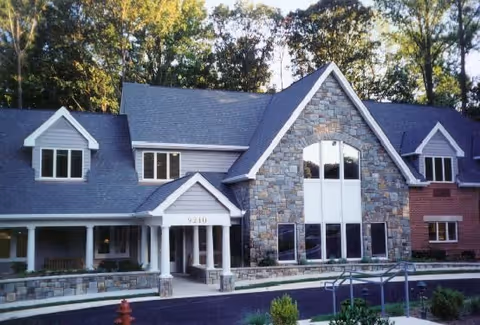 Exterior view of a two-story senior living facility building with a combination of stone, brick, and siding on the facade. The building features large windows, a peaked roof, and a covered entrance with white columns. Trees are visible in the background.