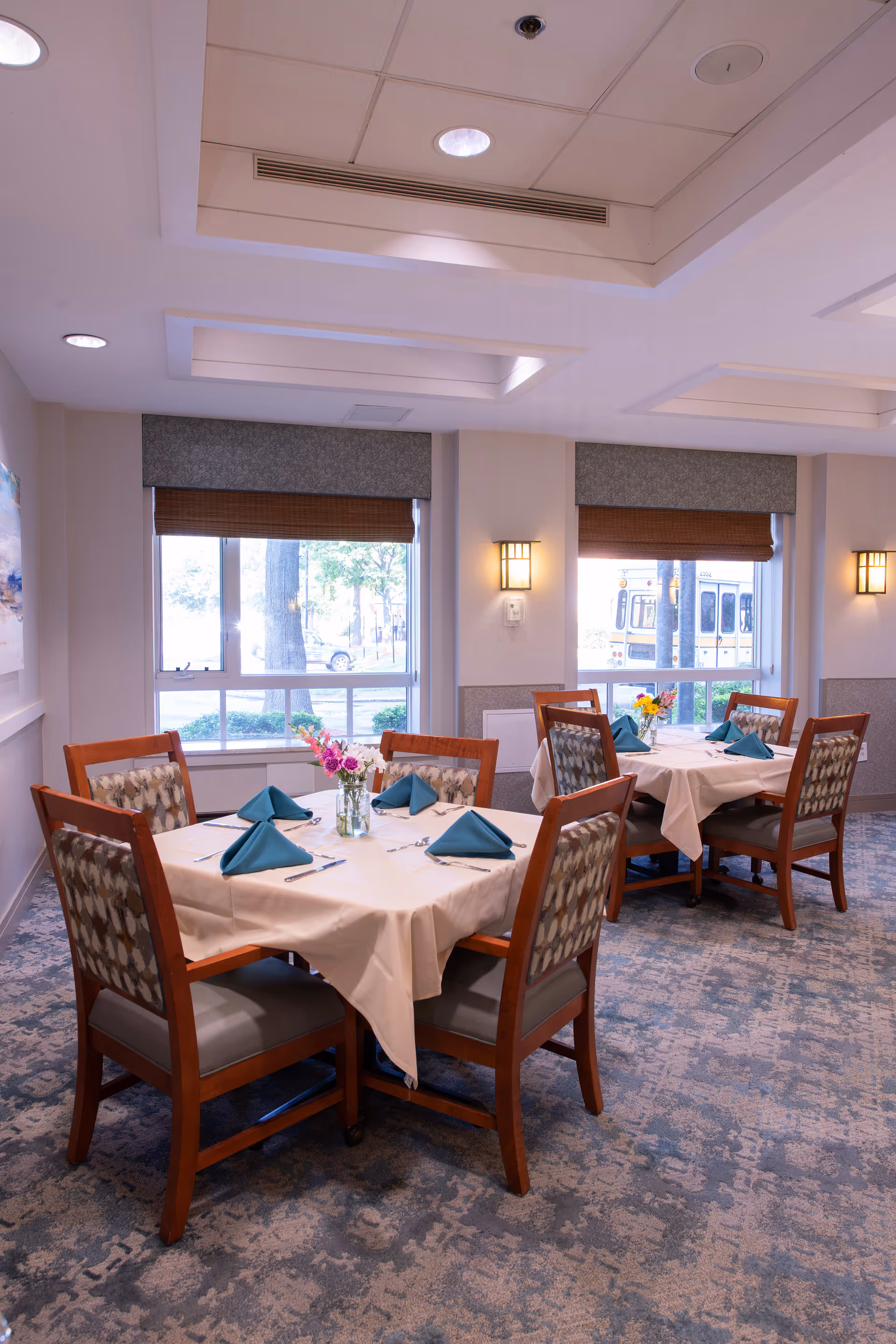 A dining room with two square tables, each set with white tablecloths, teal folded napkins, silverware, and small flower arrangements in glass vases. The room has large windows with brown blinds, patterned carpet, and soft wall lighting.