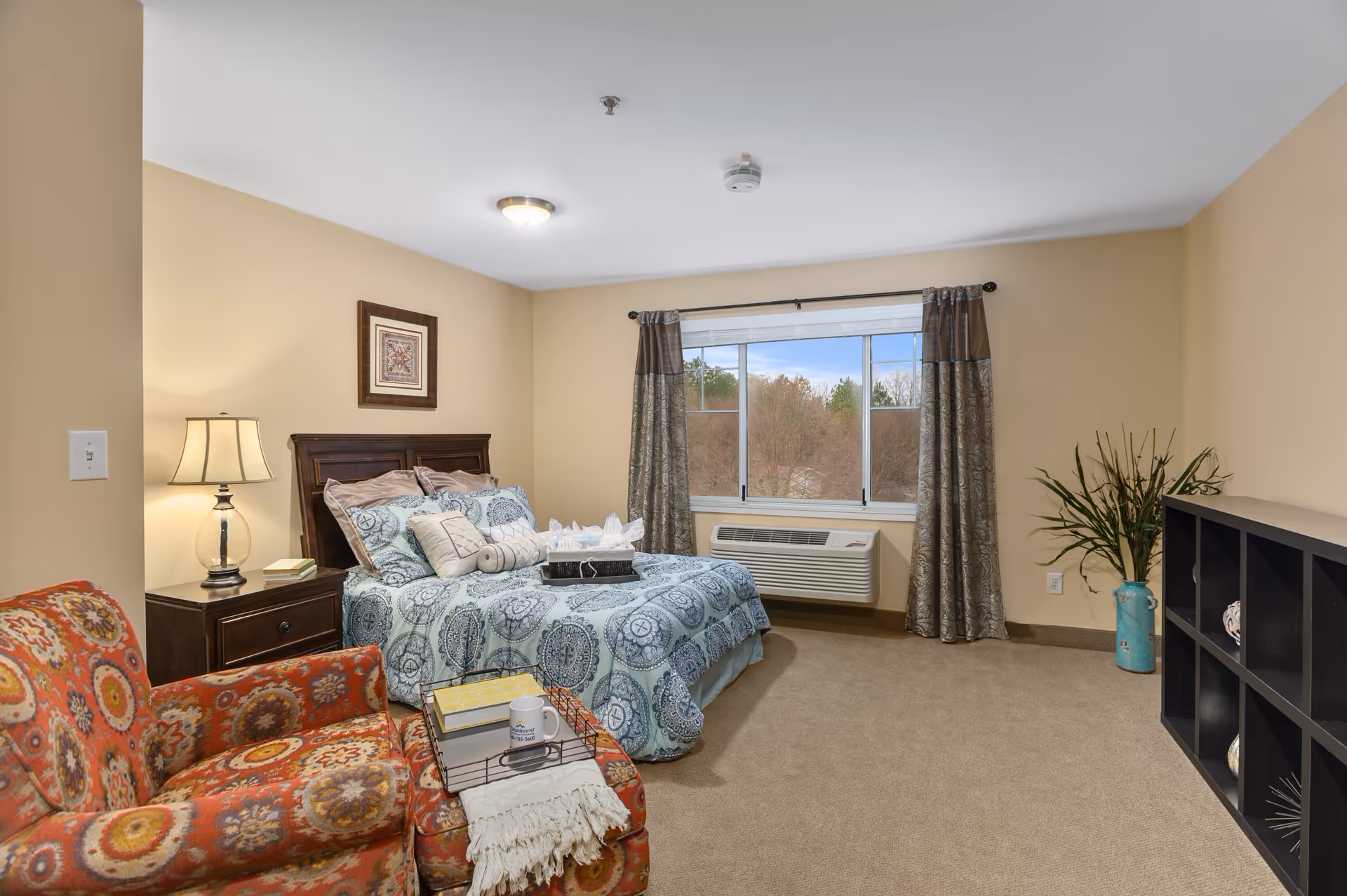 Furnished bedroom with a patterned bed and pillows, a colorful armchair, nightstand with lamp, window with curtains, and shelving.
