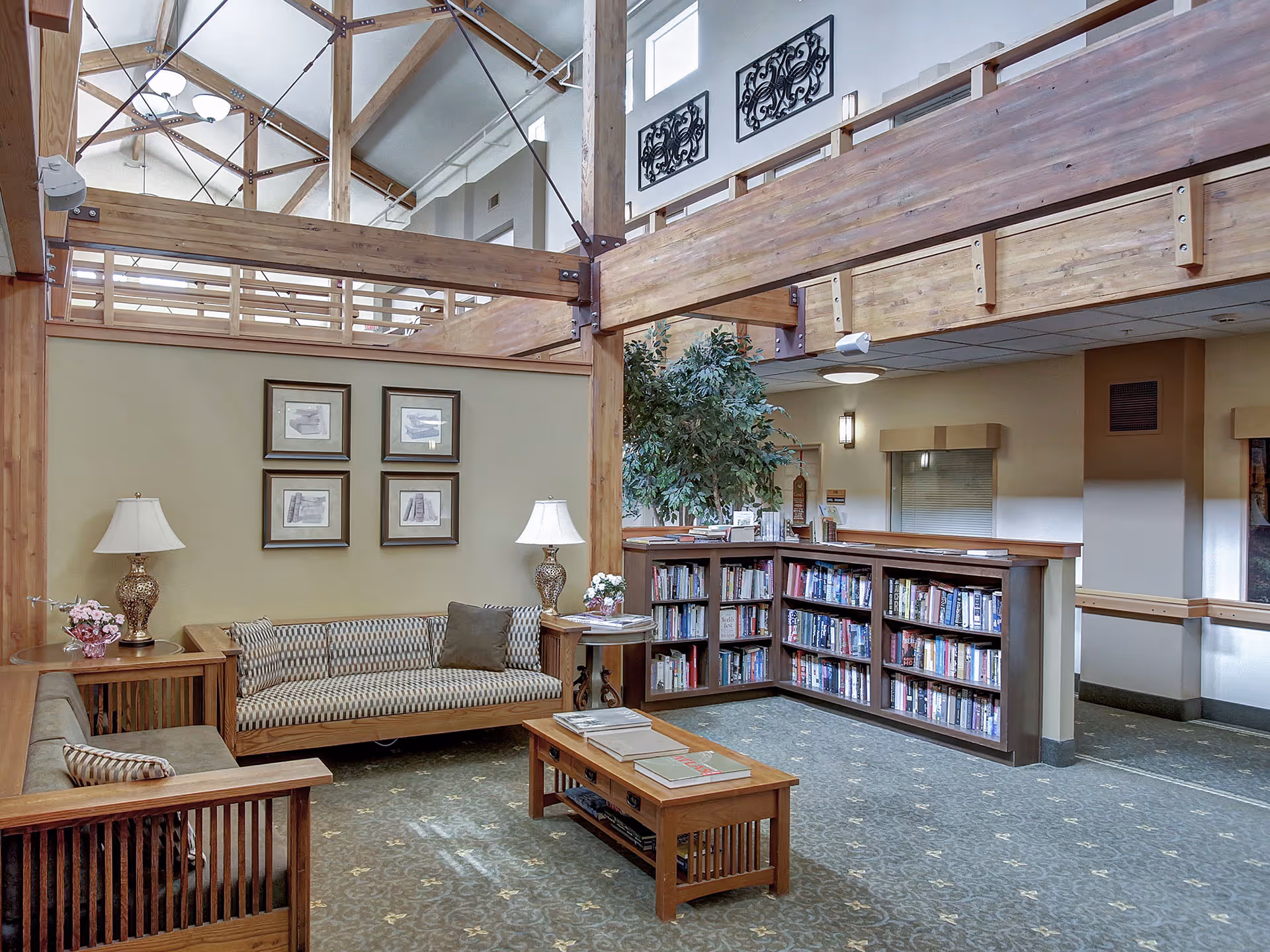 A cozy senior living common area with wooden furniture including two sofas and a coffee table. There are two decorative table lamps on side tables, a bookshelf filled with books, framed artwork on the wall, and a large indoor plant. The space features exposed wooden beams and a high ceiling with natural light coming in.