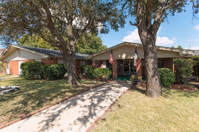 Exterior view of a single-story brick building with a covered entrance, two large trees in front, and an American flag and a Texas state flag displayed near the entrance. A concrete walkway leads up to the entrance, surrounded by grass and shrubs under a clear blue sky.