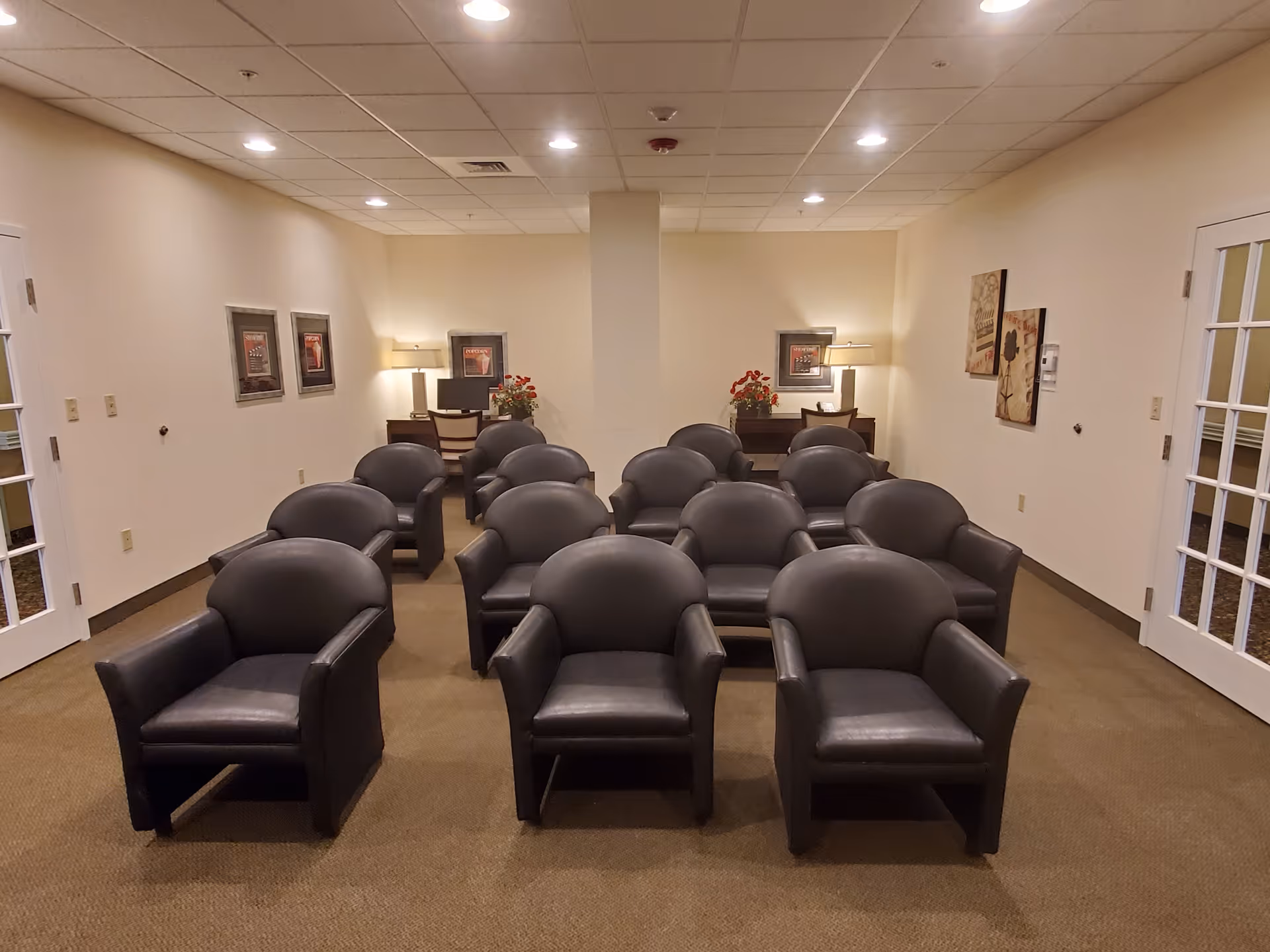 A small conference or meeting room with three rows of black leather armchairs arranged facing forward. The room has beige walls, carpeted floor, and a drop ceiling with recessed lighting. At the back of the room, there are two desks with lamps, computers, and flower arrangements. Framed pictures hang on the walls, and there are glass-paneled double doors on either side of the room.