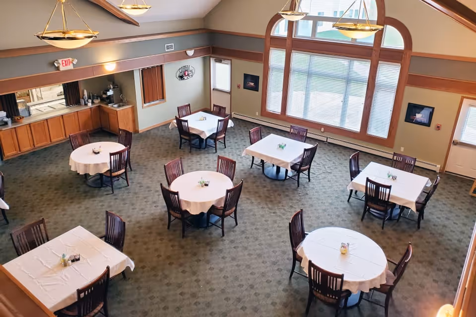 A spacious dining room with several round and square tables covered with white tablecloths, each surrounded by wooden chairs. The room features large arched windows letting in natural light, green carpeted floor, and warm-toned walls with wooden trim. There are ceiling lights and a small kitchenette area with cabinets and a counter in the corner.