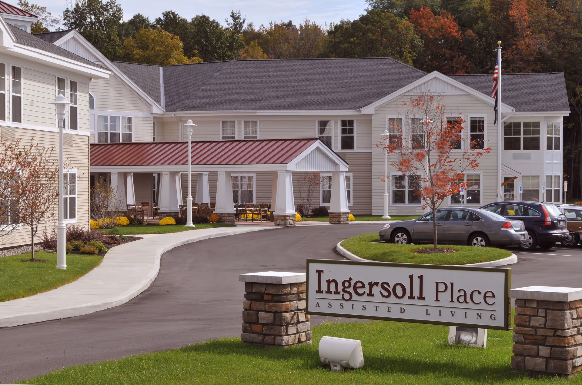 Exterior view of Ingersoll Place assisted living facility showing a two-story building with beige siding and a red metal roof over the entrance. There is a curved driveway leading to the entrance, several parked cars, a lawn with a small tree with red leaves, and an American flag on a flagpole.