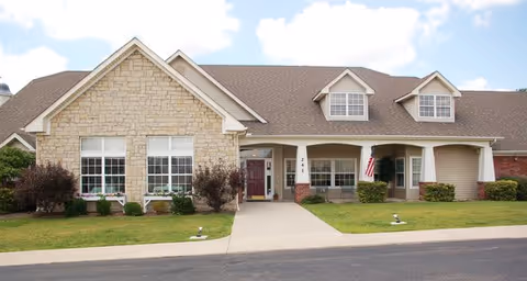 Front exterior view of a single-story senior living facility building with a stone and brick facade, multiple windows, a covered entrance with a red door, and an American flag displayed near the entrance. The building is surrounded by a well-maintained lawn and a paved driveway.