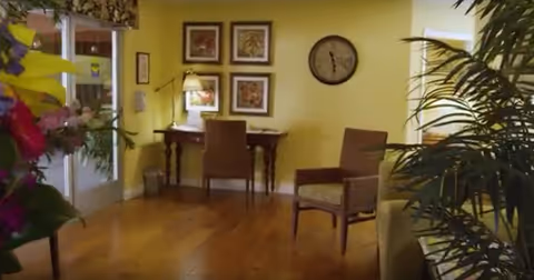 Cozy sitting area with wooden floors, a desk and chairs against a yellow wall, a wall clock and framed art, and plants by a glass door.
