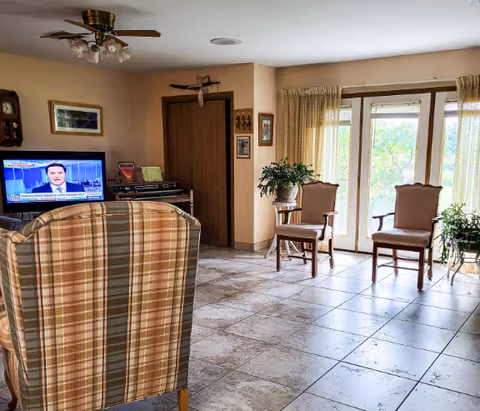 A cozy living room area with a plaid armchair facing a television mounted on the wall. The room has tiled floors, two wooden chairs near a large window with sheer curtains, and several potted plants. The walls are decorated with framed pictures and a clock.