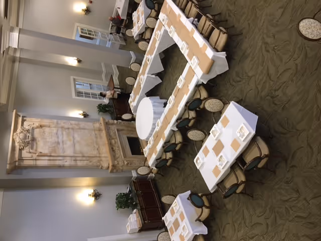 Formal dining room with tables arranged in a U-shape around a central round table, set with place settings and an ornate fireplace in the background.