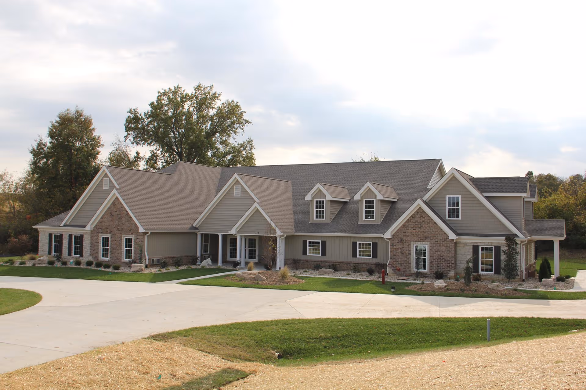 Exterior view of a large, single-story residential-style building with a combination of brick and siding, multiple gabled roofs, and several windows. The building is surrounded by a paved driveway, green lawns, and some landscaping with trees in the background under a cloudy sky.